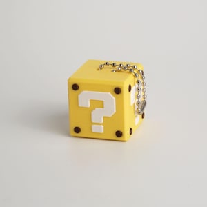 Puede incluir: Un llavero amarillo en forma de cubo con un signo de interrogación blanco en un lado y puntos marrones en las esquinas. El llavero tiene una cadena de bolas plateada.