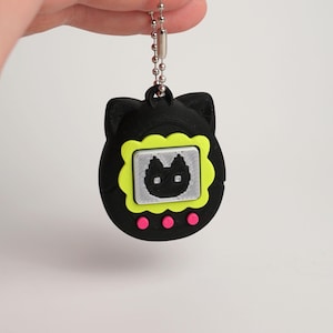 以下が含まれることがあります： 黒い猫の形をしたキーホルダー。画面にはピクセル化された猫の顔が描かれています。画面はネオンの緑色で縁取られ、下に3つのピンク色のドットがあります。キーホルダーはシルバーのボールチェーンに付いています。