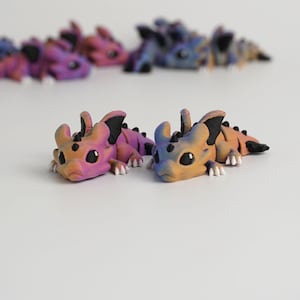 May include: Several small, colorful dragon figurines in shades of pink, purple, blue, and orange are arranged on a white surface. The dragons have black accents and white claws. The figurines are lying down.