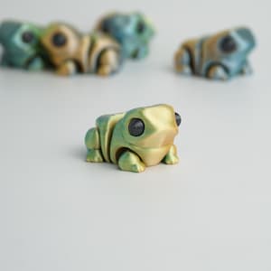 Puede incluir: Una colección de pequeñas figuras de rana impresas en 3D en tonos verdes, dorados y azules. Cada rana tiene ojos grandes y negros y una superficie texturizada. Las ranas están dispuestas sobre una superficie blanca, con una rana enfocada.