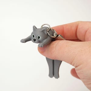 Pode incluir: Um porta-chaves em forma de gato cinzento com textura impressa em 3D, apresentando um corpo plano, patas dianteiras estendidas e um anel metálico preso ao topo da cabeça. O gato tem olhos brancos com pupilas pretas e um pequeno nariz triangular.