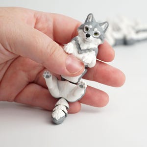 Puede incluir: Figura de lobo gris y blanco con un diseño detallado. El lobo está en una pose juguetona, con sus patas y cola visibles. La figura está sujeta por una mano sobre un fondo blanco.