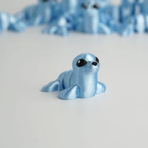 May include: A light blue seal figurine with black eyes and a black nose, lying on a white surface. Several similar figurines are blurred in the background. The seal has a smooth, slightly shiny texture.
