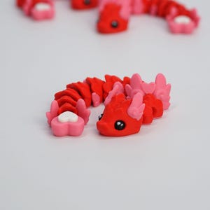 May include: A red and pink articulated dragon toy with a heart-shaped tail. The dragon has a red body, pink accents, and black eyes. Several similar toys are in the background, slightly out of focus. The toy is made of plastic.