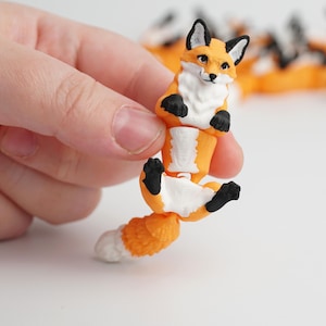 May include: A small, orange and white fox figurine with black accents. The fox has a white chest and belly, black paws and ears, and a fluffy tail. The figurine is held in a hand against a white background.