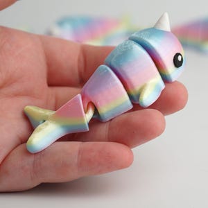 Könnte beinhalten: Ein farbenfrohes, bewegliches Narwal-Spielzeug in Pastell-Regenbogenfarben. Das Spielzeug hat ein weißes Horn, ein schwarzes Auge und einen segmentierten Körper, der Bewegung ermöglicht. Schwanz und Körperabschnitte sind in Rosa-, Gelb-, Blau- und Lilatönen bemalt.