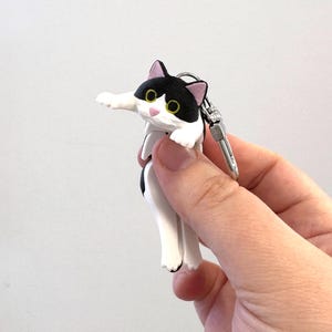 Könnte beinhalten: Schlüsselanhänger mit einer schwarz-weißen Katze, rosa Ohren und gelben Augen. Die Katze hat eine weiße Schnauze und ist an einem silberfarbenen Verschluss befestigt.