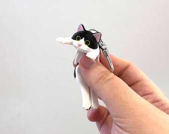May include: A black and white cat keychain with a silver clasp. The cat has pink ears, yellow eyes, and a white muzzle. The keychain is a novelty item.
