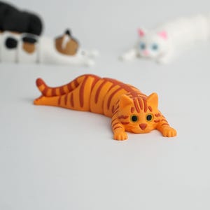 Könnte beinhalten: Eine orangefarbene getigerte Katzenfigur mit dunkelorangen Streifen, großen schwarzen Augen und einer rosa Nase, dargestellt in einer liegenden, sich streckenden Position. Zwei weitere Katzenfiguren, eine weiße und eine dreifarbige, sind im Hintergrund.