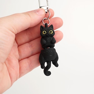 May include: Black cat keychain with yellow eyes and a silver clasp. The cat is split in two, with the top half upright and the bottom half hanging down. The keychain is made of a dark material.