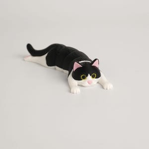 Op de afbeelding: Een zwart-witte kattenfiguur met roze oren en gele ogen. De kat ligt languit, met uitgestrekte poten. Het beeldje is een decoratief item, mogelijk voor een plank of bureau.