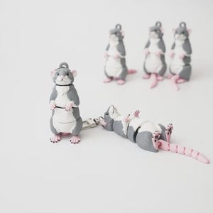 Könnte beinhalten: Eine Sammlung von fünf beweglichen Rattenfiguren, jede mit einem grau-weißen Körper sowie rosa Ohren, Pfoten und einem segmentierten Schwanz. Eine Ratte steht aufrecht, eine liegt auf der Seite und drei stehen im Hintergrund, alle mit Metallösen.