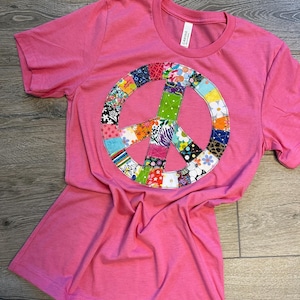 May include: A pink t-shirt with a large peace sign made of colorful patchwork fabric.