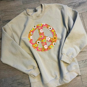May include: A light beige sweatshirt with a large peace sign made of colorful fabric with a floral pattern.