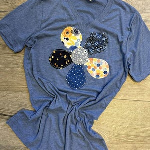 May include: A blue V-neck t-shirt with a large flower design on the front. The flower is made of various fabric patterns, including floral and polka dot designs. The shirt is made of a soft material and has short sleeves.
