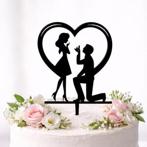 May include: A black silhouette cake topper featuring a man kneeling in a proposal pose to a woman inside a heart-shaped frame. The cake topper is decorated with pink roses and white flowers.