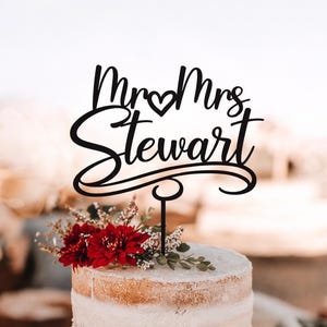 May include: A black cake topper with the words "Mr & Mrs Stewart" in a cursive font, with a heart symbol between the titles. The topper is placed on a white frosted cake decorated with red flowers and greenery.