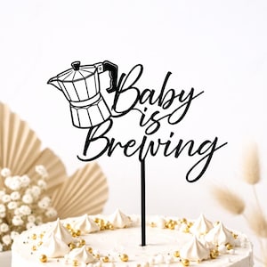 Könnte beinhalten: Schwarzer Tortenaufsatz mit der Aufschrift "Baby is Brewing" und einer Kaffeekannen-Illustration. Der Aufsatz befindet sich auf einem weißen Kuchen, der mit weißem Zuckerguss und goldenen Streuseln verziert ist. Der Hintergrund umfasst dekorative Fächer und Blumen.