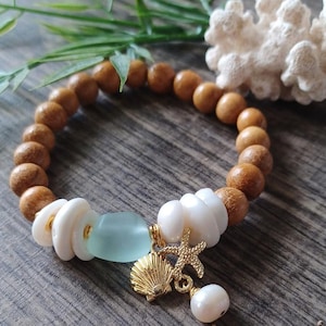 Könnte beinhalten: Ein Armband mit hellbraunen Holzperlen, weißen Muschelakzenten und einer hellblauen Glasperle. Goldene Anhänger sind eine Muschel, ein Seestern und eine hängende Perle. Das Armband hat ein Strand- oder maritimes Thema.