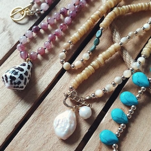 May include: A selection of handmade necklaces featuring natural materials. One necklace has a shell pendant with black markings. Other necklaces have beads in pink, beige, and turquoise. The necklaces are displayed on a wooden surface.