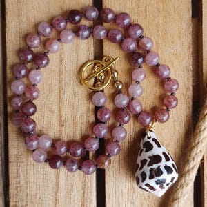 May include: A necklace with two strands of purplish-pink beads, a gold-coloured toggle clasp, and a seashell pendant with brown and white patterns. The necklace is displayed on a wooden surface. The beads are approximately 0.8 cm in diameter.
