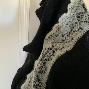 May include: A black knitted shawl with a delicate, openwork, cream-colored lace border. The shawl is draped, showcasing the contrast between the dark, solid knit and the intricate lace design. The texture of the knitwear is visible.