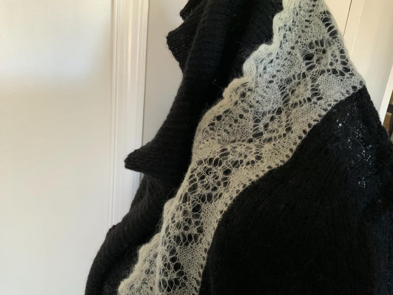May include: A black knitted shawl with a delicate, openwork, cream-colored lace border. The shawl is draped, showcasing the contrast between the dark, solid knit and the intricate lace design. The texture of the knitwear is visible.