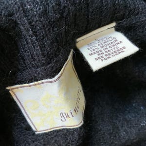 May include: Close-up of a black knit garment with two attached tags. One tag displays the brand name "guineveri" in a script font. The other tag lists the material composition: 30% nylon, 20% acrylic, and 18% mohair. The tag also indicates the item was made in China.