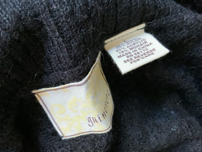 May include: Close-up of a black knit garment with two attached tags. One tag displays the brand name "guineveri" in a script font. The other tag lists the material composition: 30% nylon, 20% acrylic, and 18% mohair. The tag also indicates the item was made in China.