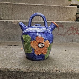 Può includere: Una brocca di ceramica blu con manico e due beccucci. La brocca è decorata con un grande fiore arancione e bianco e foglie verdi. La brocca è appoggiata su una superficie di cemento.