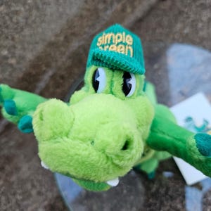 May include: A plush green alligator toy wearing a green cap with the words "Simple Green" embroidered on it. The alligator has large black eyes and white teeth. The toy is soft and cuddly.
