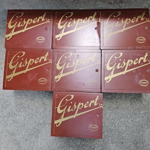 May include: A collection of seven rectangular, maroon-colored boxes. Each box features the gold-colored word "Gispert" in a cursive font. A smaller, circular emblem is also visible on each box.