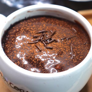 May include: A white mug filled with rich, dark chocolate pudding, dusted with cocoa powder and chocolate sprinkles. The dessert is presented in a white ceramic mug, offering a comforting treat.