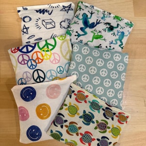 May include: A collection of small, square fabric pouches with various colorful patterns. Designs include smiley faces, peace signs, turtles, and frogs. The pouches are arranged on a light wooden surface.