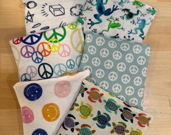May include: A collection of small, square fabric pouches with various colorful patterns. Designs include smiley faces, peace signs, turtles, and frogs. The pouches are arranged on a light wooden surface.