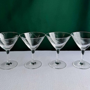 May include: Four clear glass martini glasses with etched feather designs. The glasses have a classic triangular bowl shape, slender stems, and round bases. The background is a dark green color, and the glasses are arranged on a white surface.