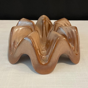 May include: A ceramic, wave-shaped ashtray in shades of brown and tan. The ashtray has a glossy finish and a unique, geometric design. The ridges create multiple resting spots for cigarettes or cigars. The piece is likely vintage.