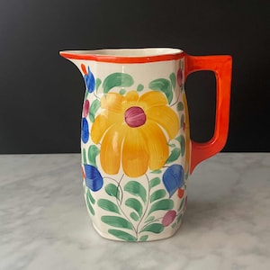 May include: A ceramic pitcher with a vibrant floral design. The pitcher features a large yellow flower with a purple center, surrounded by green leaves and blue and red accents. It has an orange handle and rim.
