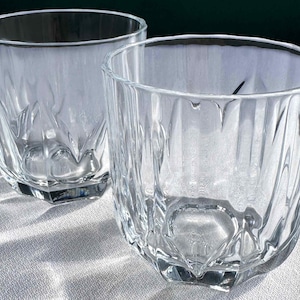 May include: Two clear, faceted drinking glasses. The glasses have a thick, geometric base and a textured, vertical pattern on the sides. The glasses are empty and appear to be made of crystal or high-quality glass.