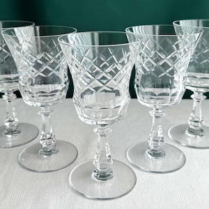 May include: Five clear crystal wine glasses with diamond-cut patterns. Each glass has a flared bowl, a slender stem, and a round base. The glasses are arranged on a white surface against a dark green background.