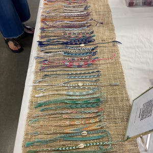 May include: A display of handmade friendship bracelets in various colors and patterns. The bracelets are arranged on a woven runner, showcasing a variety of designs, including some with small shell accents. The color palette ranges from vibrant pinks and oranges to cool blues and greens.