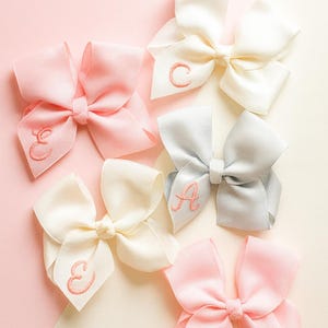Pode incluir: Uma coleção de laços de cabelo feitos à mão em várias cores, incluindo rosa, branco e cinza. Cada laço apresenta uma inicial bordada em uma cor correspondente. Dispostos sobre um fundo bicolor rosa e creme.