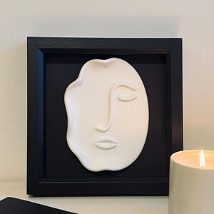 Op de afbeelding: Een witte abstracte gezichtssculptuur in een zwarte schaduwbox. De sculptuur heeft een minimalistisch ontwerp met een enkel oog, neus en mond. Een brandende kaars en een zwart notitieboek met pen staan op de voorgrond.