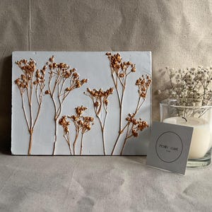 May include: A rectangular, light blue resin art piece with embedded dried flowers in shades of gold and brown. A clear glass candle holder with a lit white candle and a sprig of white flowers sits to the right. A small card with the text "resin cast" is in front.
