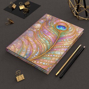 May include: A decorative journal featuring a colorful peacock feather design with gold accents and jewel-like embellishments. The journal is surrounded by gold office supplies, including paper clips and pencils, on a dark gray surface.