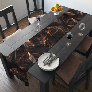 May include: A dining table set for a meal, featuring a black table runner with a geometric design in shades of black and copper. The table is set with plates, silverware, and wine glasses. A bowl of fruit sits at the table's center.