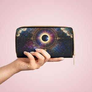 Pode incluir: Uma carteira preta com um design celestial com um brilho solar central e um eclipse escuro. A carteira tem um zíper dourado e um puxador a condizer. O fundo é uma mistura de azuis, roxos e rosas, assemelhando-se a uma galáxia.
