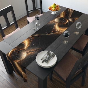 May include: A black dining table is set with a decorative table runner featuring a gold and black celestial design. The table is set with wine glasses, plates, and silverware. A bowl of fruit sits at the top of the table.