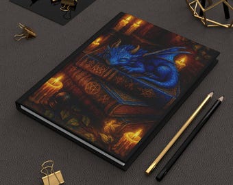 May include: A black journal featuring a detailed illustration of a sleeping blue dragon atop a stack of books, surrounded by lit candles. Gold and black pencils and gold binder clips are also visible.