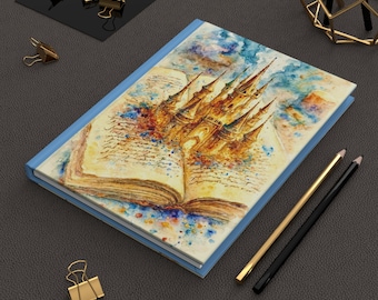 May include: A hardcover journal with a light blue cover and a watercolor illustration of a castle emerging from an open book. The artwork features warm tones of gold, brown, and blue. Gold and black pencils and gold binder clips are nearby.